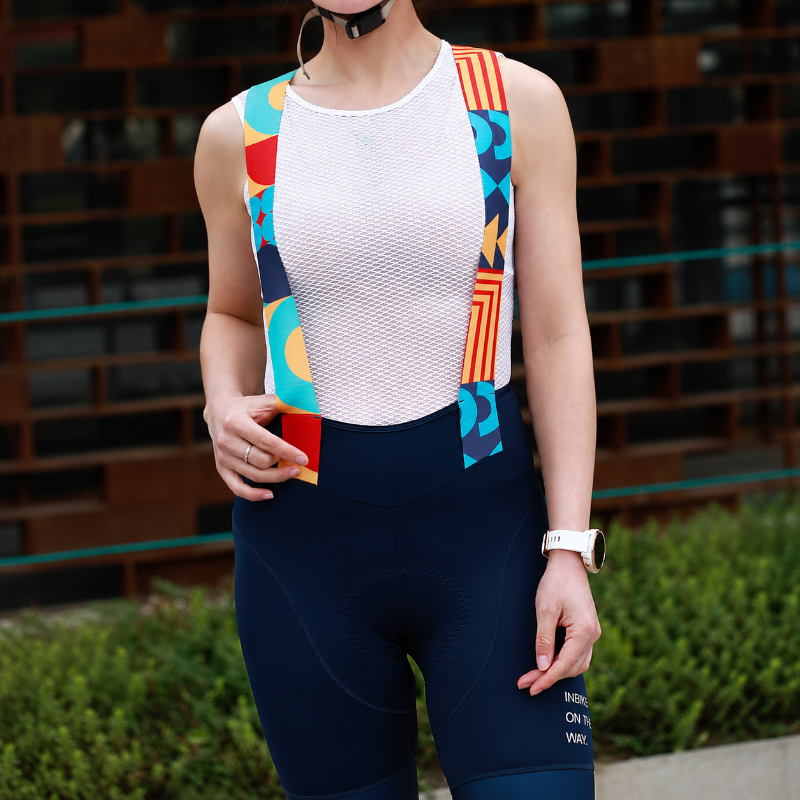 A woman wearing a white mesh cycling jersey with colorful geometric patterned shoulder panels and navy blue cycling shorts, standing outdoors in front of a brick building.