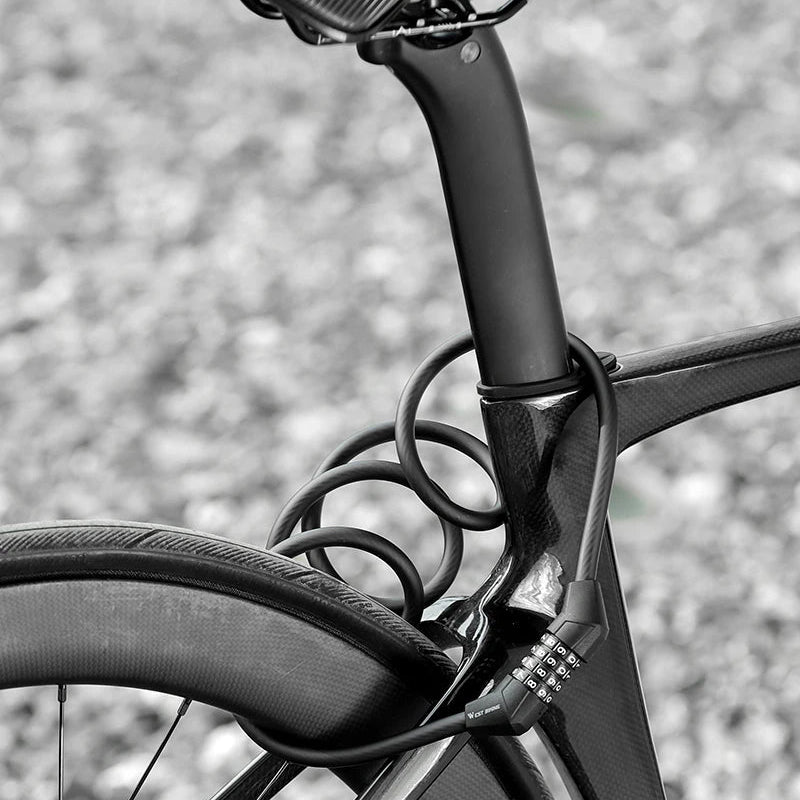 A close-up black and white photograph of a bicycle frame and saddle, showing a coiled lock around the seat post area. The background is a blurred gravel surface.