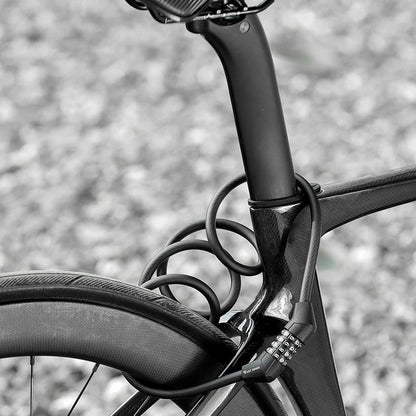 A close-up black and white photograph of a bicycle frame and saddle, showing a coiled lock around the seat post area. The background is a blurred gravel surface.