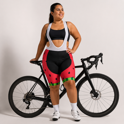 A woman stands next to a black road bicycle wearing a black and white cycling bib top and red watermelon-print cycling shorts with green rind details. She is posing in front of a light gray studio background.