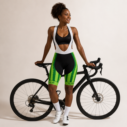 A woman stands next to a black road bike wearing a black and white cycling sports bra and matching black and green bib shorts. The shorts feature green vertical stripes and green side panels