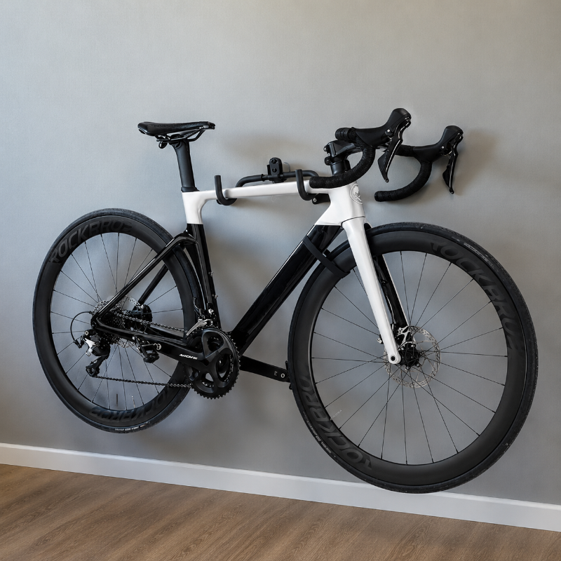 A black and white road bicycle with drop handlebars, mounted against a light gray wall on a wooden floor.
