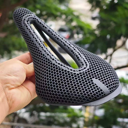 A black bicycle saddle with a honeycomb mesh pattern, held by a hand against a blurred outdoor background of green trees.