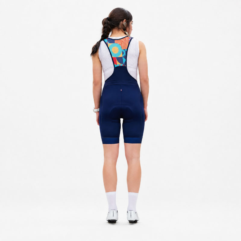 A woman viewed from behind wearing a navy blue cycling bib shorts set with white shoulder panels and a colorful geometric pattern on the back, paired with white socks and cycling shoes on a white studio background.