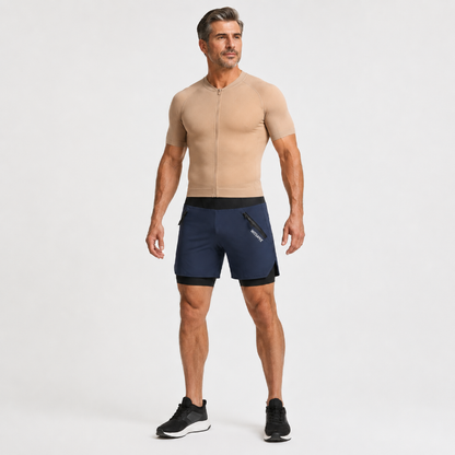 A man modeling a beige short-sleeve zip-up compression shirt and navy blue athletic shorts with black trim, standing against a light gray studio background.