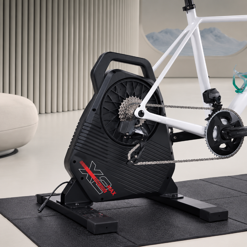 A black indoor cycling trainer with a white bicycle mounted on it, placed on a dark mat in a modern indoor setting with a curved white wall and a light-colored floor.