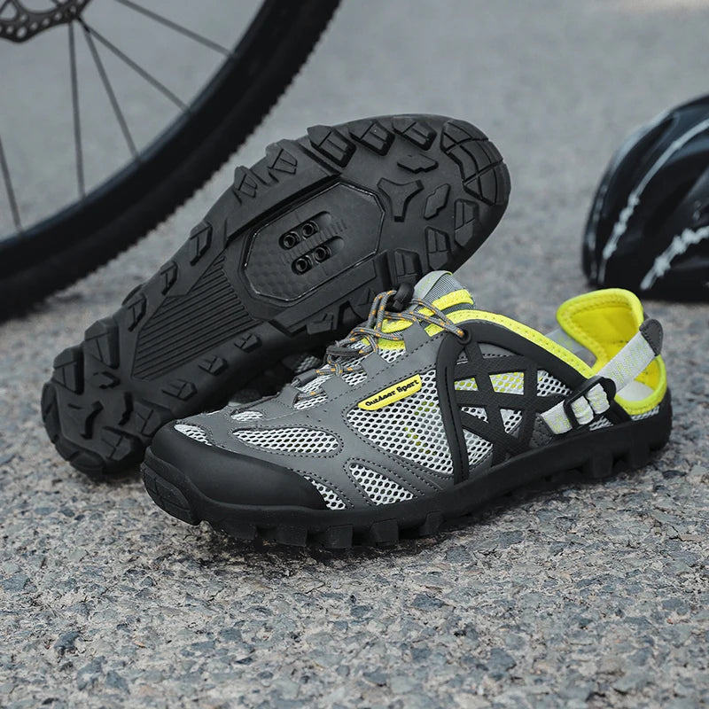 A pair of gray and black cycling shoes with bright yellow accents, displayed on a gravel surface with a bicycle wheel in the background.