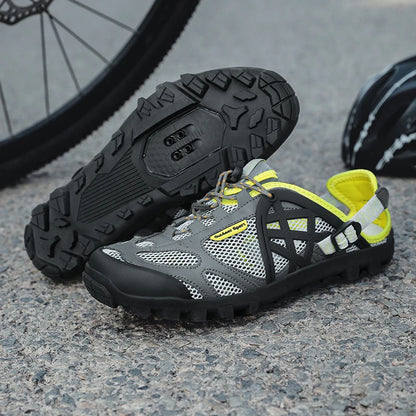 A pair of gray and black cycling shoes with bright yellow accents, displayed on a gravel surface with a bicycle wheel in the background.