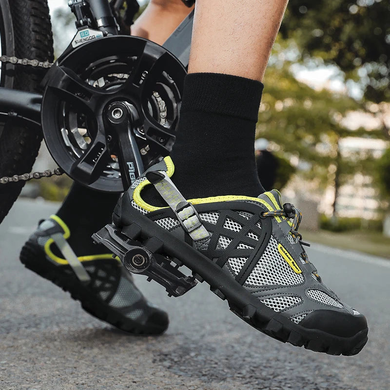 A close-up lifestyle shot of a cyclist's feet wearing gray and black cycling shoes with neon yellow accents, mounted on a bicycle crankset outdoors on a paved path.