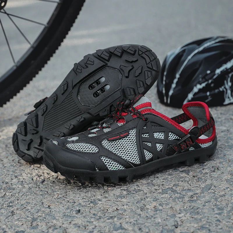 A pair of black and gray cycling shoes with red accents, shown on a gravel surface with a bicycle wheel and helmet in the background.