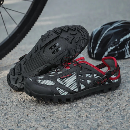 A pair of black and gray cycling shoes with red accents, shown on a gravel surface with a bicycle wheel and helmet in the background.