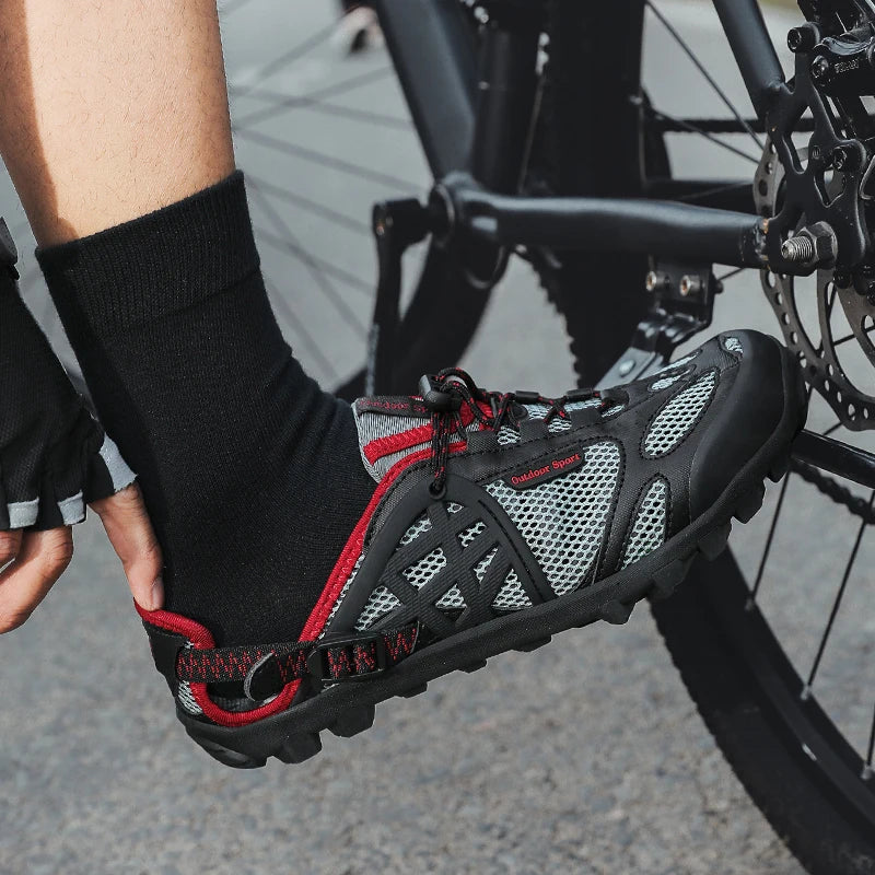 A close-up of a person's foot wearing a black and gray cycling shoe with red accents, resting on a bicycle near the rear wheel.