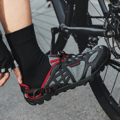 A close-up of a person's foot wearing a black and gray cycling shoe with red accents, resting on a bicycle near the rear wheel.