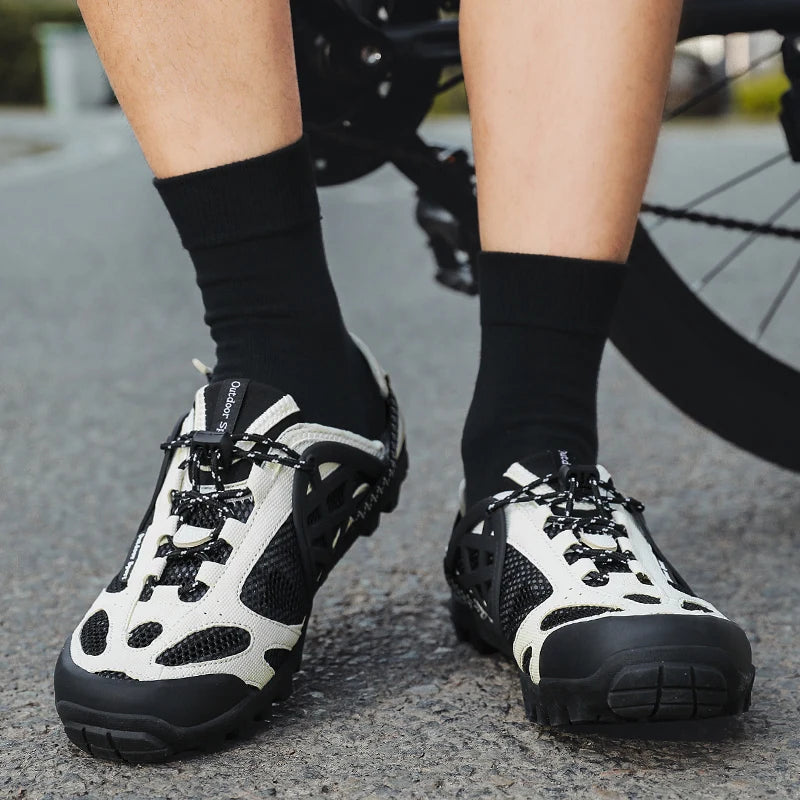 A close-up of a person's legs wearing black and off-white cycling shoes with black socks, standing on asphalt next to a bicycle.