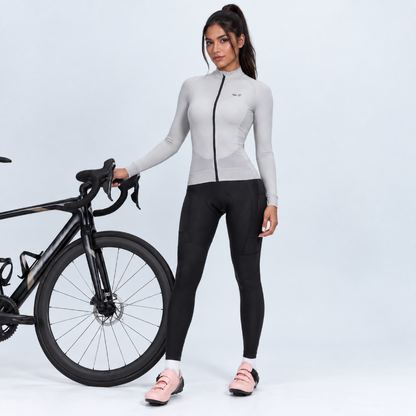 A woman stands next to a black road bicycle wearing a light gray long-sleeve cycling jersey with a black front zipper and black cycling tights. She is wearing light pink cycling shoes against a light gray studio background.