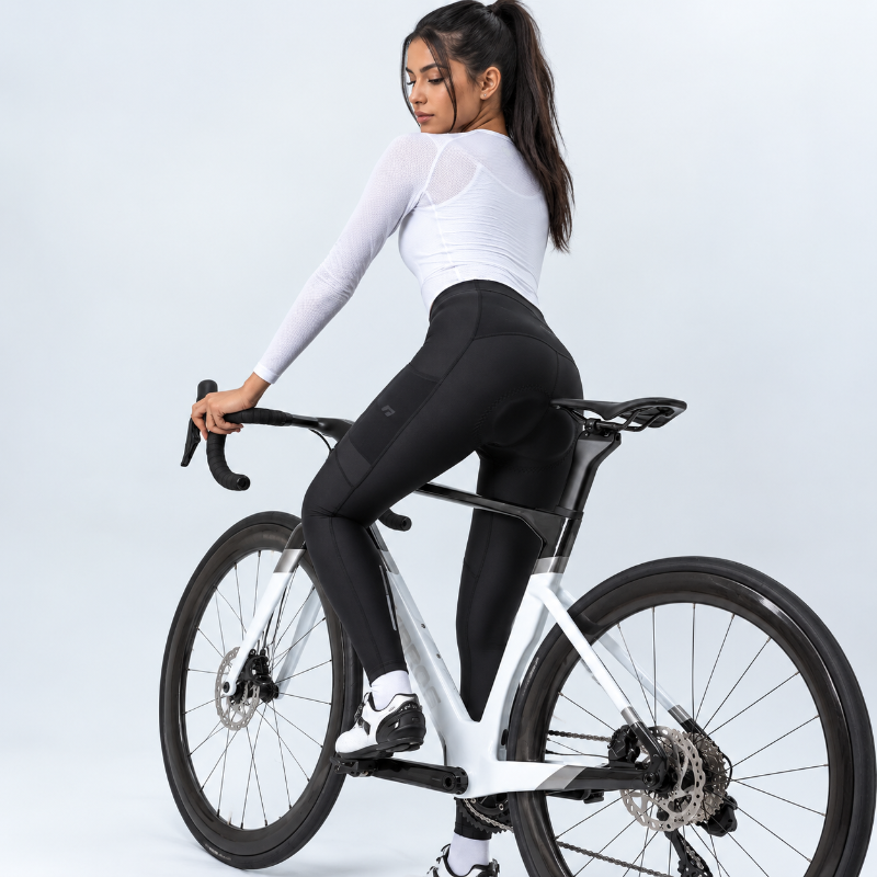 A woman wearing a fitted white long-sleeve top and black cycling tights rides a white road bicycle against a clean light gray studio background.