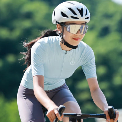 A woman riding a road bicycle outdoors, wearing a white cycling helmet, mirrored sunglasses, a light blue short-sleeve cycling jersey, and dark gray fitted shorts. The background is a blurred green natural setting.