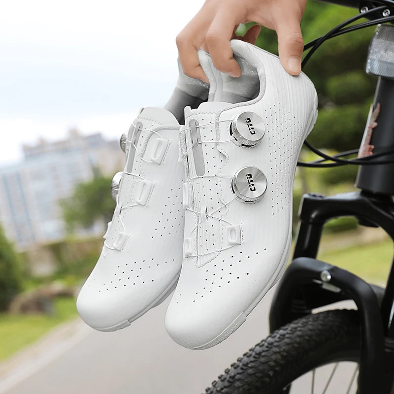 White cycling shoes with cleats held by a person next to a bicycle on a road.