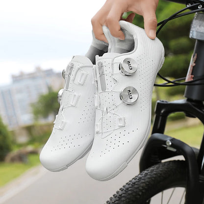 White cycling shoes with cleats held by a person next to a bicycle on a road.