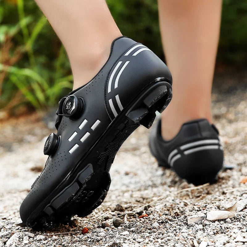 Black cycling shoes with dial closures worn on a person's feet outdoors on a gravel path with green foliage in the background.