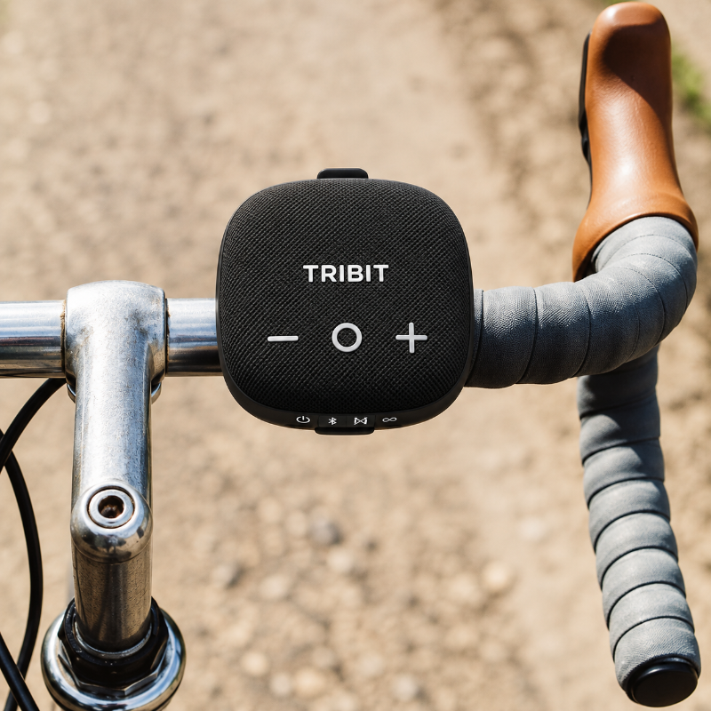 A black Tribit portable Bluetooth speaker mounted on a bicycle handlebar, with a brown leather grip and gray tape on the handlebar bar ends, set against a blurred outdoor background.